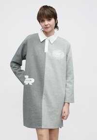 Pilka džemperio suknelė su balta apykakle. Dizainas pasižymi suskirstyta pilka spalvų schema, logotipo spauda ir ilgomis rankovėmis. Minkšta audinio tekstūra.