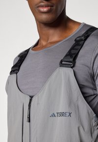 Gray waterproof bib overall with adjustable black straps and a zippered front, featuring the "TERREX" logo. Worn over a gray long-sleeve shirt.