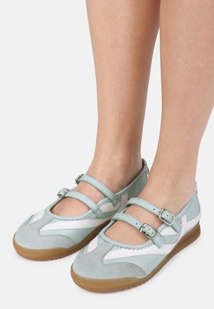 Pieds portant des chaussures style Mary Jane bleu pâle et blanc avec double bride à boucle et semelles en caoutchouc sur un fond uni.