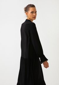 Musta pitkähiha dressi, jossa on levenevä helma, valmistettu sileästä kankaasta. Värityksessä on rypytetty yksityiskohta vyötäröllä ja frilla hihansuissa.