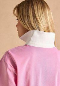 Ružový bavlnený top s bielym vysokým golierom, hladkou štruktúrou a voľným strihom, zobrazený zozadu, pričom zdôrazňuje dizajn goliera a vlasy.