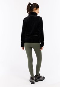 Schwarze Fleecejacke mit hohem Kragen, kombiniert mit olivgrünen Rippleggings und schwarzen Wanderschuhen mit silbernen Akzenten.