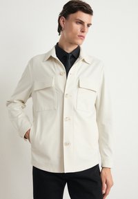 Lys beige jakke med krage, to frontlommer og knappelukking. Laget av glatt stoff, kombinert med en svart skjorte og slips.