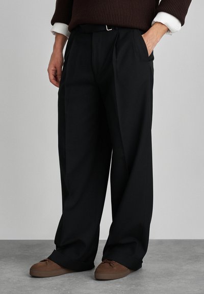 Black wide-leg trousers made of smooth fabric, featuring front pleats, side pockets, and a silver buckle on the waistband. Brown shoes visible.