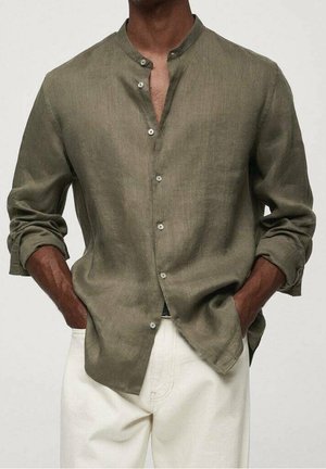 Homme portant une chemise vert olive à col mandarin boutonnée, manches retroussées, et un pantalon blanc, mains dans les poches.