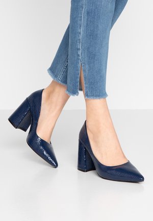 Mørkeblå patenterede læder højhælede sko med en struktureret overflade, spids tå og kraftig hæl, parret med jeans med frynset kant.