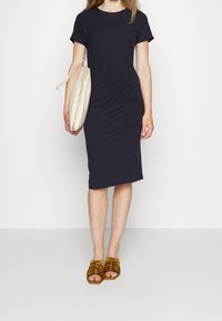 Marinblå klänning med kort ärm och rundad rynkig sidodetalj. I kombination med en beige handväska och gula sandaler med remmar. Slät textur.