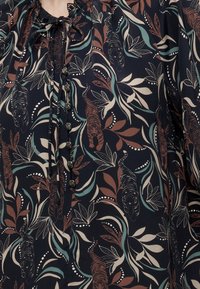 Blouse foncée avec motifs tigre et feuilles multicolores, devant boutonné et détail noué au niveau de l'encolure.
