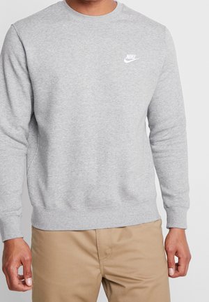 Mężczyzna w jasnoszarej bluzie Nike i beżowych spodniach, stojący z rękami swobodnie opuszczonymi na tle jednolitego, jasnego tła.