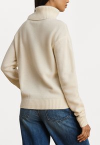 Pull de gola alta creme feito de um tecido suave, com um colarinho e punhos canelados, apresentando um ajuste reto e relaxado. Usado com jeans de ganga azuis.