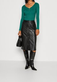 Haut vert côtelé à manches longues avec un col en V et un détail de fermeture éclair dorée, associé à une jupe midi en cuir noir avec des boutons et des bottes noires montant jusqu'aux genoux.