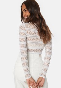Top bianco a maniche lunghe in pizzo con un design a motivi, caratterizzato da una silhouette aderente e maniche testurizzate, che mostrano dettagli intricati.