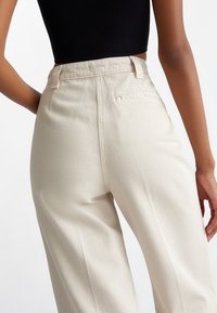 Beige højtaljede bukser lavet af bomuld, med lige ben design, baglomme og en glat tekstureret overflade.