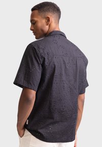 Black short-sleeve shirt with intricate cut-out patterns, featuring a collar and textured fabric. Back view shows design details and relaxed fit.
