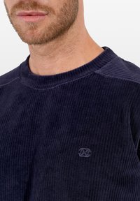 Granatowa bluza sztruksowa o prążkowanej fakturze i okrągłym dekolcie. Na piersi znajduje się haftowane logo.