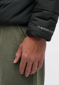 Črna izolirana jakna z gladko teksturo, ki ima vidno oznako "THERMA-FIT" na rokavu; v kombinaciji z olivno zelenimi tkaninastimi hlačami.