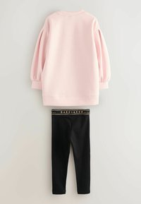 Lichtroze langemouwtop met pofmouwen, gecombineerd met zwarte leggings met een gouden "BAKER" logo op de tailleband.