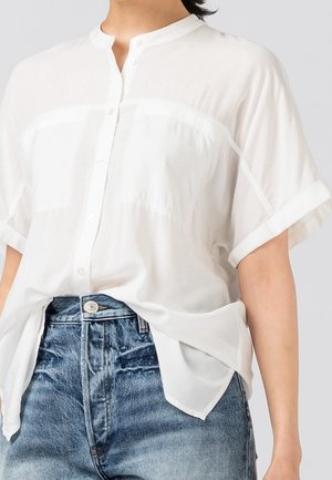 Femme portant une chemise blanche à manches courtes et ample, rentrée légèrement dans un jean bleu taille haute avec une fermeture à bouton visible.