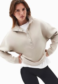 Jasno beżowy sweter z wysokim kołnierzem, z zapięciem na guziki i miękką teksturą. Noszony na białej koszuli i zestawiony z czarnymi legginsami.