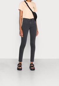 Camiseta melocotón con un gráfico de girasol, bolso bandolera negro, jeans ajustados oscuros y sandalias negras con suelas gruesas. Tela suave y líneas limpias.