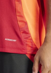 Maglietta atletica rossa e arancione, con texture in mesh, cuciture di design curvato e marchio AEROREADY. Pantaloni corti neri visibili alla base.