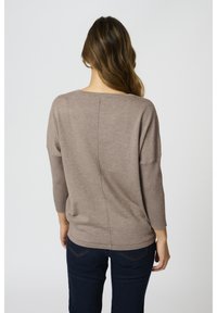 Brązowy sweter dzianinowy o luźnym kroju, z rękawami do trzech czwartej długości, o prążkowanej strukturze i środkowym szwie na plecach. Noszony z ciemnymi dżinsami.