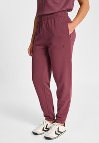 Bordowe spodnie dresowe z elastycznym pasem, bocznymi kieszeniami, zwężanymi mankietami i subtelnym logo marki. Nosi się je z czarno-białymi sportowymi butami.