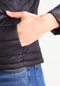 Sort puffjakke med quiltet design, der har en sidelomme og en glat, shiny overflade. Kombineret med blå denim jeans.