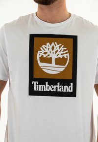 T-shirt bianco a maniche corte con una grafica centrale che presenta un quadrato marrone con un logo ad albero bianco e la parola "Timberland" sotto in grassetto nero.