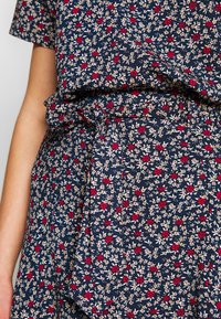 Tissu bleu marine avec un petit motif floral en rouge, blanc et beige. Présente un design ample avec un détail de taille nouée.