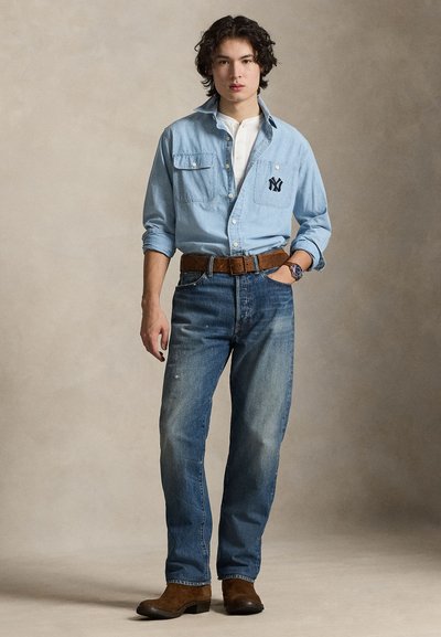 Svetlomodra denim srajca z dvema prsnikoma in logotipom NY, v kombinaciji z modrimi kavbojkami in rjavimi usnjenimi škornji ter rjavim pasom z teksturo.