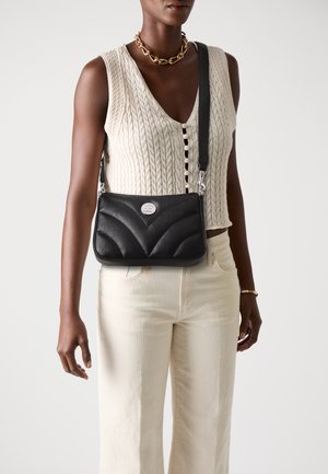 Borsa a spalla in pelle nera con design a zigzag trapuntato, hardware argentato e tracolla regolabile, abbinata a un top in maglia color crema e pantaloni chiari.