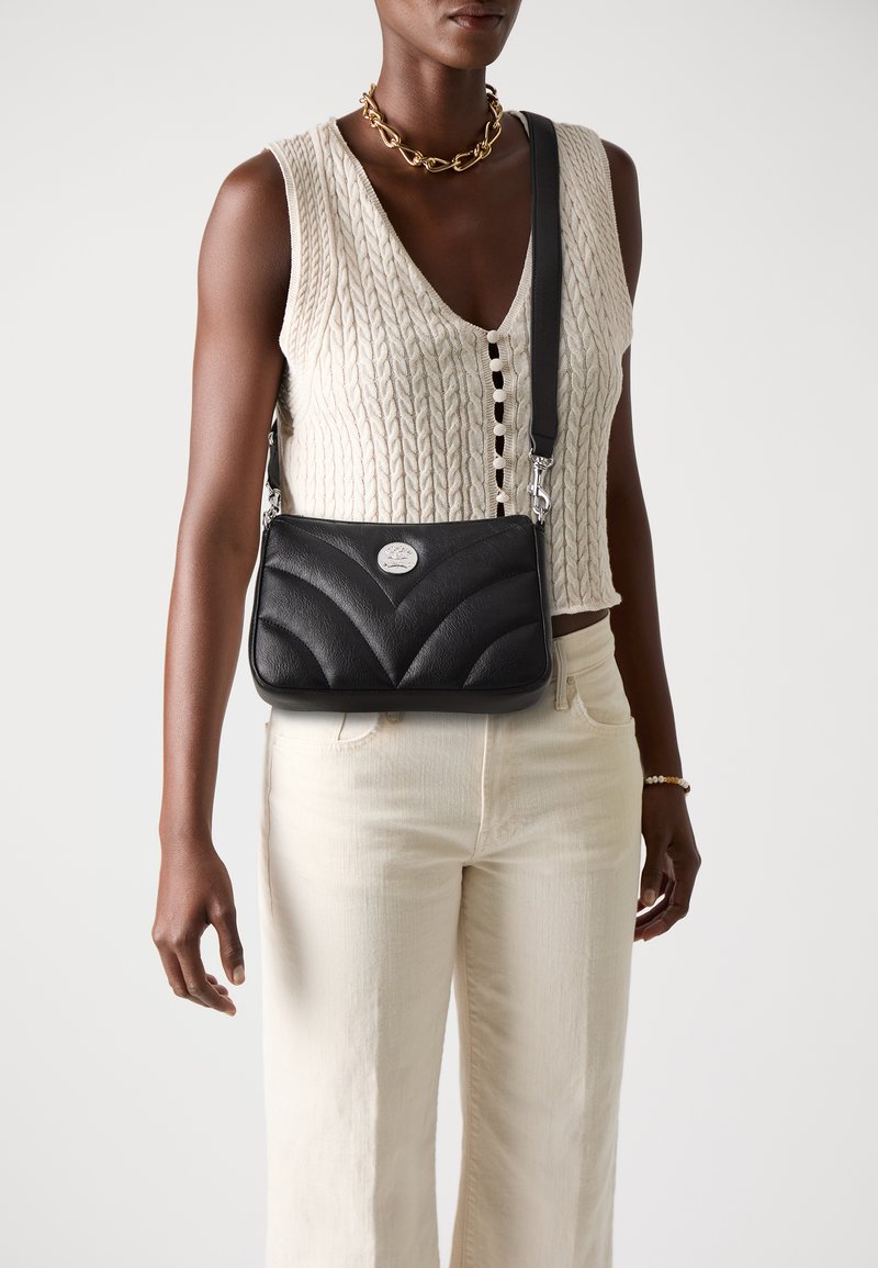 Borsa a spalla in pelle nera con design a zigzag trapuntato, hardware argentato e tracolla regolabile, abbinata a un top in maglia color crema e pantaloni chiari.