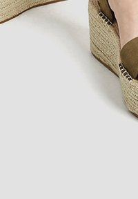 Espadrille à talon compensé avec une plateforme en jute tissé et un dessus en tissu vert olive doux. Présente des accents en métal argenté sur les côtés.