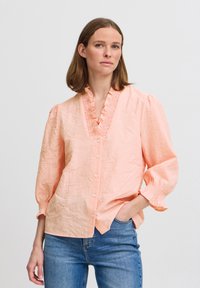 Peach-colored blouse with a textured surface, ruffled neckline, and puffed sleeves. Features front button closure and relaxed fit.
