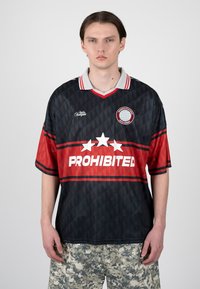 Joven con pelo liso y oscuro lleva una camiseta deportiva negra y roja con el texto "PROHIBITED" y estrellas, combinada con pantalones cortos de camuflaje gris.