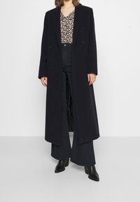 Cappotto lungo doppiopetto in lana blu scuro, con classici rever a punta e tasche frontali, indossato sopra una blusa floreale e pantaloni a gamba larga.