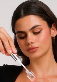 Vrouw met donker haar brengt een transparante lipgloss aan met een applicator, draagt een zwart shirt en kleine oorringen, neutrale achtergrond.