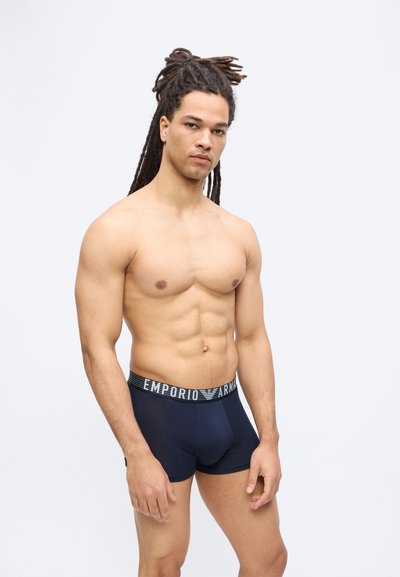 Μυώδης άνδρας με μακριές κοτσίδες ντρέντλοκ, φορώντας ναυτικό μπλε ανδρικά εσώρουχα Emporio Armani, σε λευκό απλό φόντο.