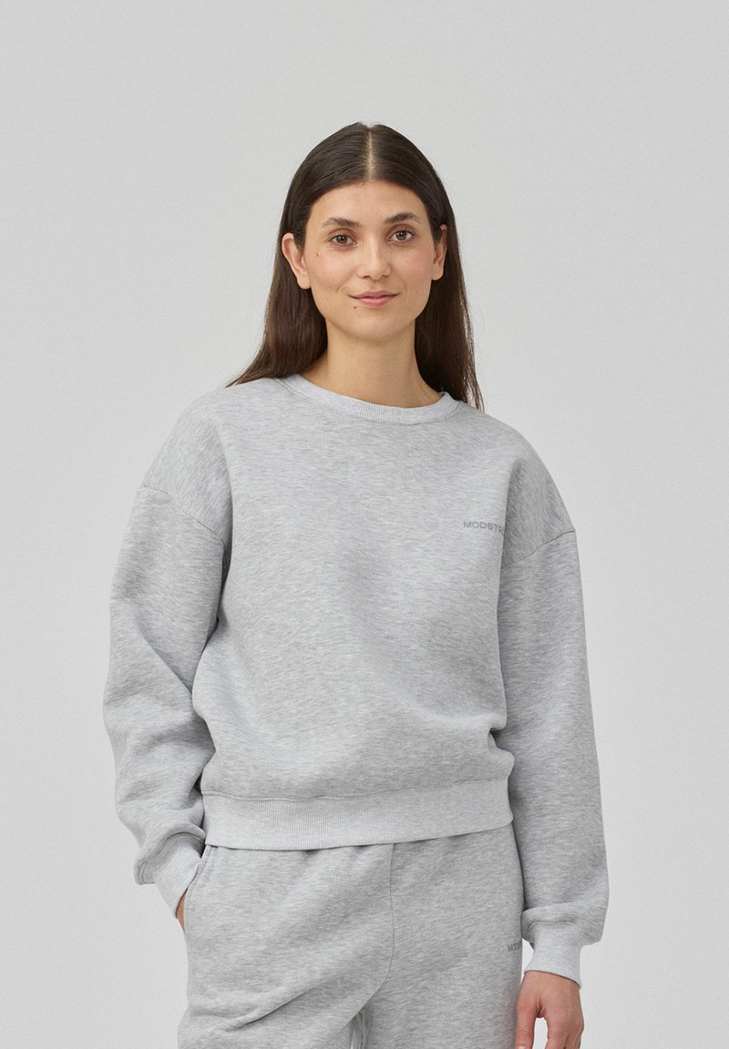 Sweatshirt gris en tissu doux, avec un col rond, des épaules tombantes et des poignets côtelés. Comprend un détail de logo discret sur la poitrine.