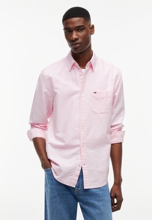 Νεαρός άνδρας που φοράει ροζ ριγέ πουκάμισο με κουμπιά και γυρισμένα μανίκια, και μπλε τζιν, στέκεται με το ένα χέρι στην τσέπη.