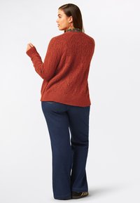 Rostfärgad stickad tröja med en texturerad mönster, i kombination med mörkblå utsvängda jeans. Tröjan har lång ärm och rundad halsringning.