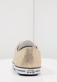 Zapatilla de lona texturizada dorada con suela de goma blanca, rayas negras y logo en el talón. Diseño de parte trasera redondeada y de corte bajo.