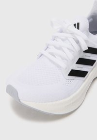Valge atleetjalats, millel on kootud ülemine osa, kolm musta triipu, tekstureeritud kangas ja heledat värvi vaheline tald, millel on "LIGHT BOOST" bränding.
