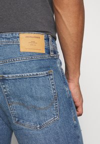 Jeans de ganga azul com um padrão desgastado. Apresentam um rótulo de pele bege na cintura traseira e costura amarela nos bolsos.