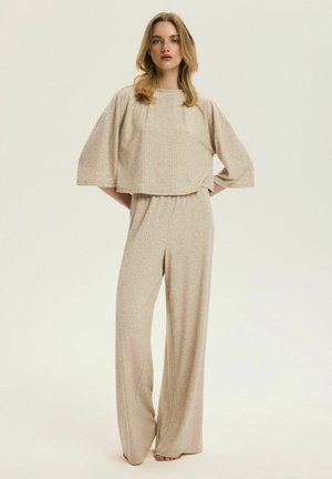 Kobieta stojąca boso, ubrana w beżowe, szerokie spodnie z prążkowanego materiału oraz pasujący luźny top z krótkim rękawem, na jednolitym tle.