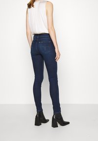 Štíhlé tmavě modré džíny s vysokým pasem, které mají zadní kapsy a žlutou logovou nášivku, spárované s černými kotníkovými botami na blokovém podpatku.