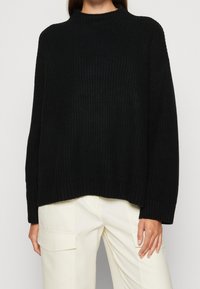 Schwarzer Rippstrickpullover mit hohem Kragen, oversized Passform und überschnittenen Schultern, kombiniert mit cremefarbenen Cargo-Hosen.