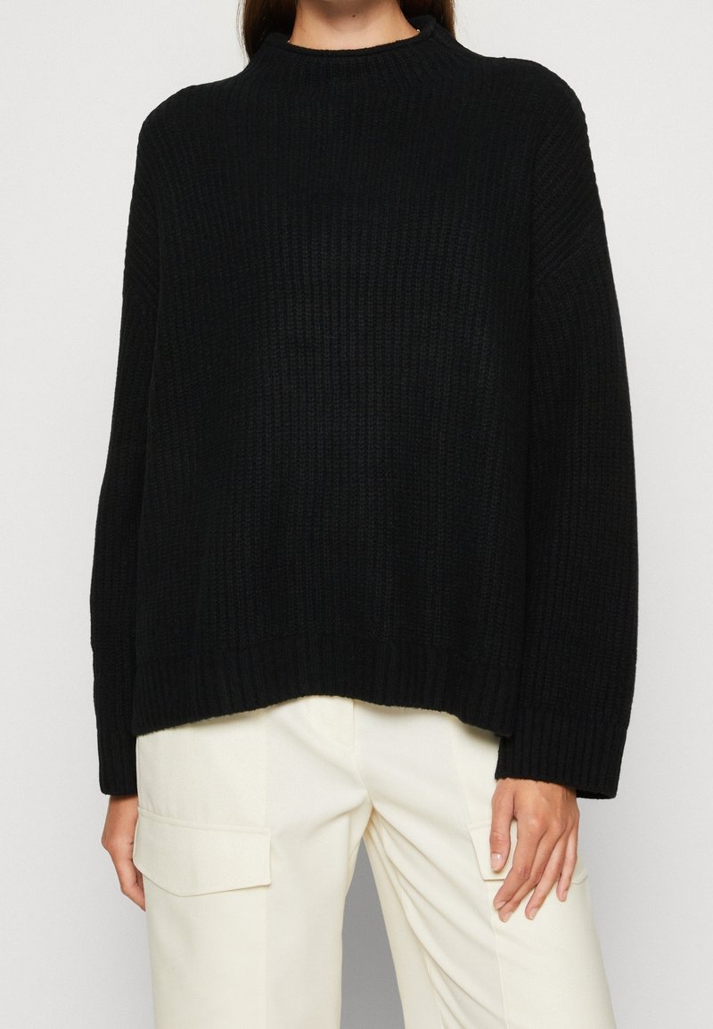 Schwarzer Rippstrickpullover mit hohem Kragen, oversized Passform und überschnittenen Schultern, kombiniert mit cremefarbenen Cargo-Hosen.