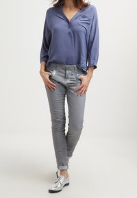 Světle modrá košile s výstřihem do V a jednou kapsou, kombinovaná se šedými slim-fit kalhotami se modrým prošitím. Stříbrné šněrovací boty.